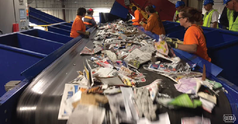 Don't go wishcycling - be sure what you put in the bin is safe to recycle [Image: workers sorting items on a conveyor belt at a recycling center] | Iowa DNR